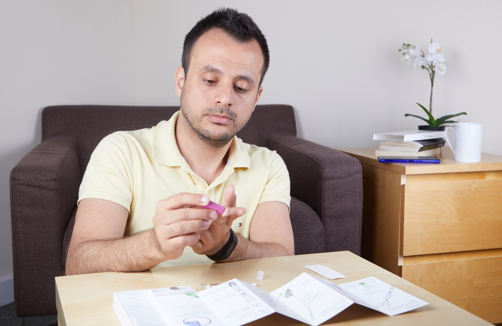 A man pricks his finger so that he can test himself for HIV. In front of him are the instructions for the test. He is concentrating on pricking his finger. The image has a pink and purple filter over it. 