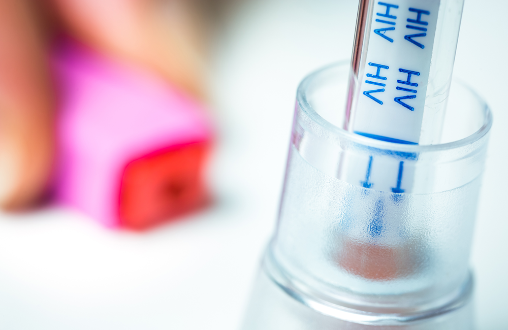 A close-up of a glass receptacle which contains a testing vial. This vial is labelled as being ready to receive blood for an HIV test. The image has a pink and purple filter over it. The image has a pink and purple filter over it. 