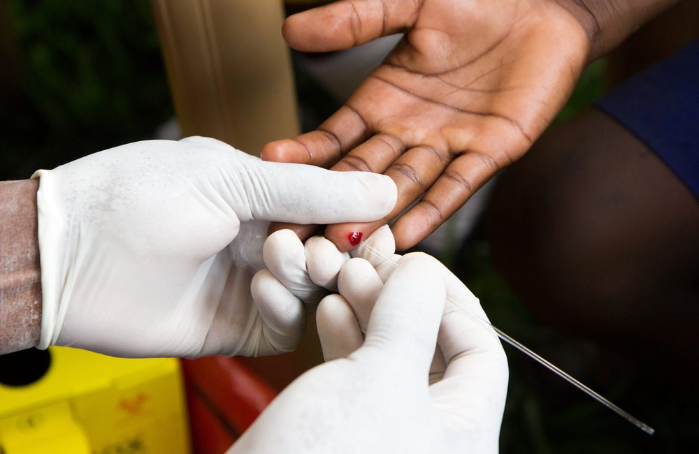 A person wearing a surgical glove holds the hand of somebody else. This person is having their finger pricked with a fine needle. The needle has drawn a small amount of blood which is visible. The image has a pink and purple filter over it. 