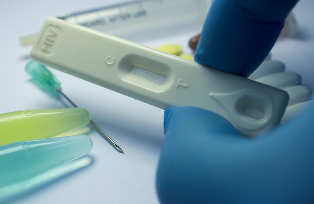 A close-up image of somebody holding an HIV test between two of their fingers. The test has not been taken yet and is blank. The person is wearing blue surgical gloves. The image has a pink and purple filter over it. 