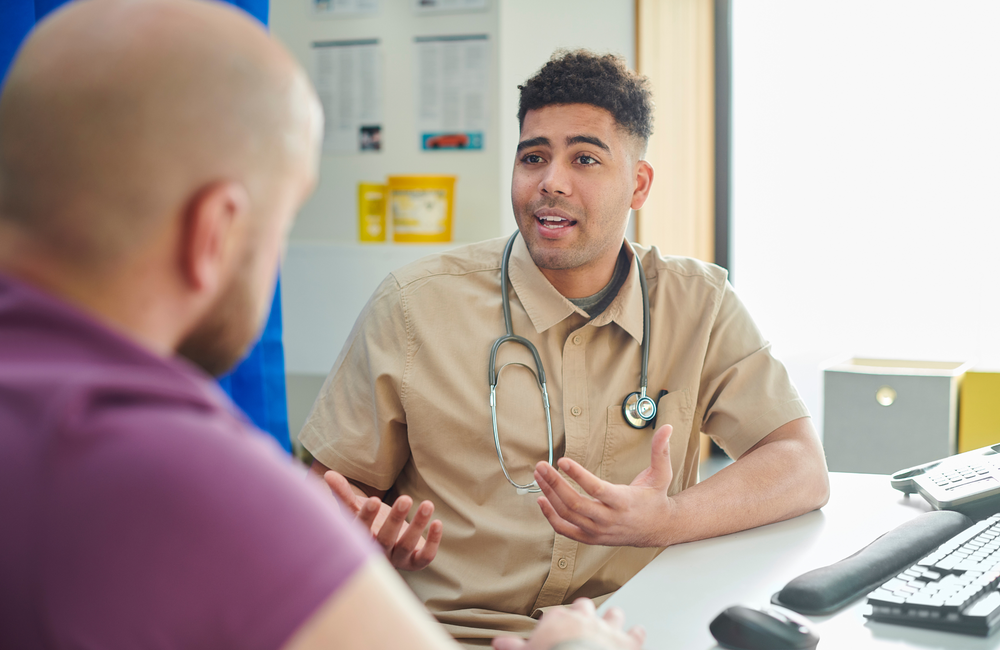 iStock A doctor wearing a light brown shirt and a stethoscope around his neck sitting at a desk and talking to a patient who's wearing a purple t-shirt.